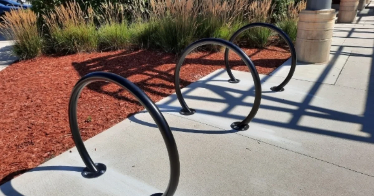 Circular black metal bike racks installed on a concrete sidewalk near landscaped greenery.